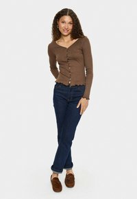 Vrouw met krullend haar glimlacht terwijl ze een bruine knooptrui, donkerblauwe jeans en bruine instappers draagt, met gekruiste benen staand tegen een witte achtergrond.