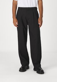 Man som bär lös svarta plisserade byxor, svarta kraftiga skor och en vit skjorta, stående mot en enkel ljus bakgrund.