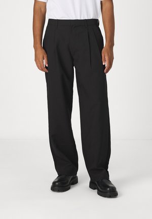 Hombre con pantalones sueltos negros con pliegues, zapatos negros gruesos y una camisa blanca, de pie contra un fondo claro y liso.