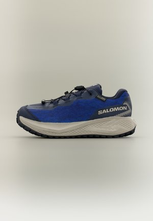 Modro-šedá bežecká topánka Salomon trail s hrubou textúrovanou podrážkou a systémom rýchleho sťahovania šnúriek zobrazená z boku na jednofarebnom pozadí.