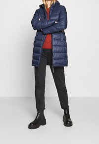 Marineblaues Puffer-Jacke mit Kapuze, mit gestepptem Design und Reißverschluss. Kombiniert mit einem roten Strickpullover und schwarzen Jeans, akzentuiert durch schwarze Plateaustiefel.