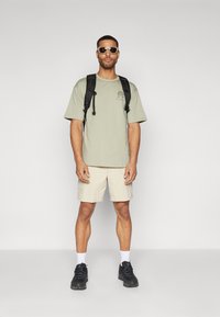 Lys grøn t-shirt med logo, beige shorts, sort rygsæk, sorte sko og hvide sokker. Solbriller fuldender det afslappede udendørs look.