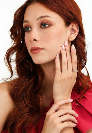 Young woman with long red hair and natural makeup touches her face, wearing a diamond ring and a red off-shoulder top against a white background.