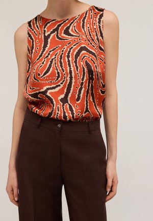 Oranssi ja musta abstraktilla kuviolla varustettu hihaton paita yhdistettynä tummanruskeisiin korkeavyötäröisiin housuihin, esitetty kaulasta lantioon.