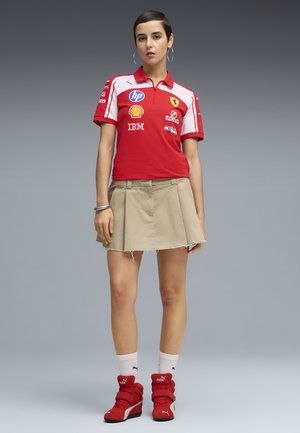 Jeune femme aux cheveux courts portant une chemise de course Ferrari rouge, une jupe beige, des chaussettes blanches Puma et des chaussures de course rouges Puma, debout devant un fond gris.