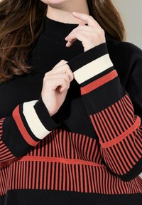 Schwarzer Pullover mit gerippter Textur, hohem Kragen und gestreiften roten und cremigen Akzenten an den Ärmeln. Enthält ausgeprägte horizontale und vertikale Muster.