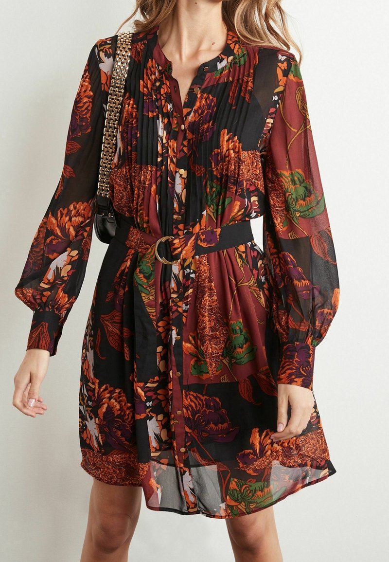Femme portant une robe transparente à manches longues à motifs floraux dans des tons noirs, orange, verts et rouges, avec une taille ceinturée et un sac en bandoulière à chaîne dorée.