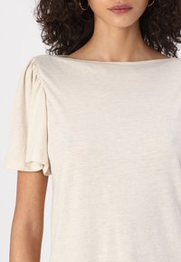 Kreminės spalvos trumpų rankovių palaidinė su pūstomis rankovėmis, apvaliu kaklu ir lengvu, tekstūruotu audiniu. Nėra matomų raštų ar akcentų.