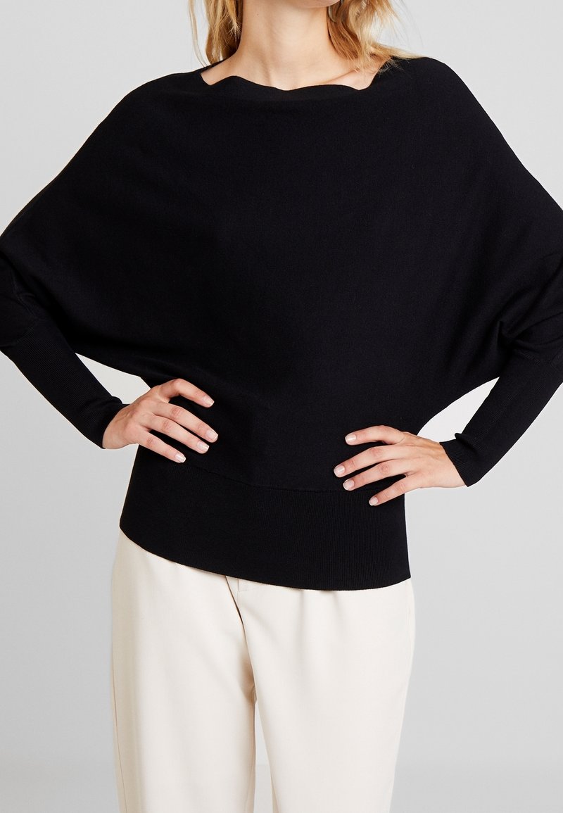 Femme portant un pull ample noir à manches longues larges avec des poignets ajustés, associé à un pantalon beige clair, mains sur les hanches.