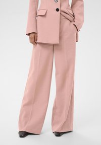 Helles rosa maßgeschneidertes Anzugensemble bestehend aus einem langen Blazer mit Reverskragen und weiten Hosen. Glatter Stoff mit dezenter Textur. Schwarze Knöpfe als Akzent.