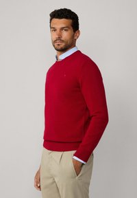 Homme portant un pull rouge à col rond sur une chemise à col bleu clair et un pantalon beige, se tenant avec une main dans la poche contre un fond uni.