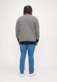 Veste à motifs noir et blanc avec des poignets et un ourlet côtelés, associée à un jean bleu et des baskets blanches. Le mannequin est vu de dos.