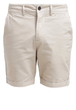 Homme portant un short beige retroussé, des chaussures habillées noires et une chemise à motif noir et blanc, les mains dans les poches.