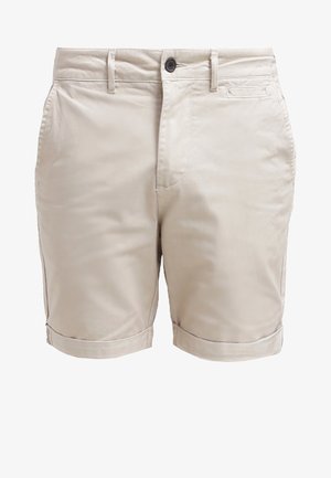 Homme portant un short beige retroussé, des chaussures habillées noires et une chemise à motif noir et blanc, les mains dans les poches.