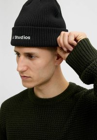 Czarna dzianinowa czapka beanie z podwiniętym brzegiem, z białym napisem "J.J. Studios". Teksturowana powierzchnia o dopasowanym kroju. Zielony sweter z prążkowanym kołnierzem.