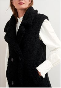 Mustat, hihattomat liivit, jotka on valmistettu teksturoidusta fleece-kankaasta. Niissä on leveä kaulus ja kaksi nappia edessä. Päällä valkoinen paita.