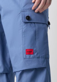 Calças cargo azul claro com um tecido texturizado, apresentando um bolso lateral com botões pretos e um emblema vermelho da HUGO.