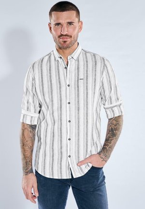 Mann mit kurzen Haaren und Armtätowierungen, der ein weißes und graues, vertikal gestreiftes Hemd mit hochgekrempelten Ärmeln trägt und mit einer Hand in der Tasche steht.