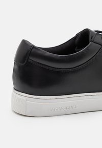 Black leather sneaker with round toe, smooth finish, white rubber sole, and textured accent. Features branding along the side.