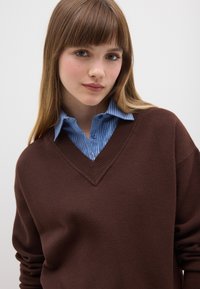 Maglione marrone a scollo a V indossato sopra una camicia a righe azzurro chiaro con bottoni. Il maglione presenta una scollatura a coste e una vestibilità ampia. Texture morbida visibile.