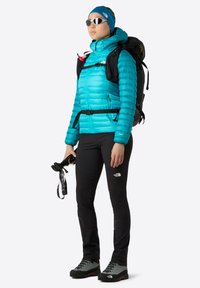 Ljusblå isolerad jacka, svarta byxor, grå vandringsskor, solglasögon, svart ryggsäck. Plagget har dragkedjor, remmar och en huva.