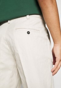Pantalon beige clair avec une finition texturée, doté d'une poche arrière avec un bouton noir. Porté avec une chemise vert foncé.