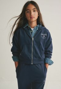 Felpa con zip color navy con tasche anteriori, abbinata a una camicia di denim. Presenta polsini a costine e logo del marchio ricamato sul petto.