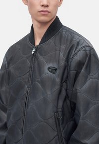 Jeune personne portant une veste matelassée gris foncé avec fermeture éclair et col côtelé, légèrement de profil sur un fond uni.