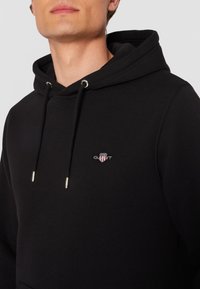 Melns džemperis ar kapuci un sudrabotiem acuļiem, uz krūtīm maza izšūta logotips. Izgatavots no mīksta auduma.