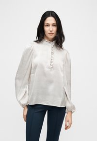 Γυναίκα με σκούρα μαλλιά που φοράει κρεμ μακρυμάνικο μπλουζάκι με λεπτές κάθετες ρίγες και γιακά με βολάν, συνδυασμένο με σκούρο εφαρμοστό παντελόνι.