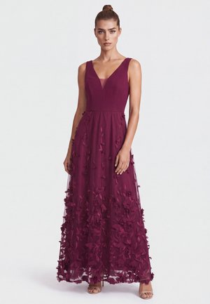 Woman with hair in a bun wearing a sleeveless deep purple gown with floral appliques and nude heels, standing against a plain light background.