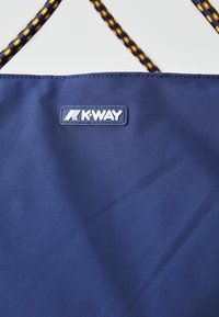 Tote en tissu bleu marine avec une texture lisse, portant un patch logo K-WAY. Les anses sont ornées d'un design tissé aux motifs jaune, orange et noir.