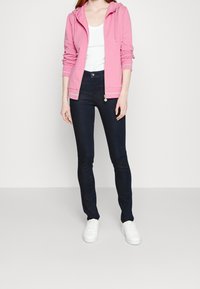 Rosa zip-up hoodie med snörning, bärs över en vit linne, i kombination med mörkblå tajta jeans och vita sneakers.