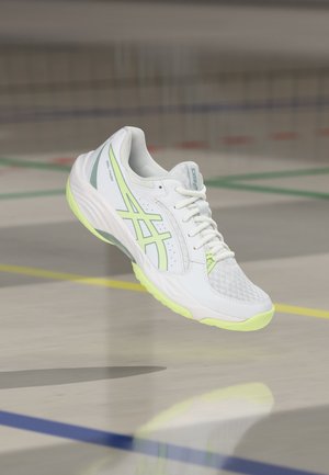 Hvid og neon gul Asics sportssko i luften over en indendørs sportsbane med farvede grænselinjer synlige på gulvet.
