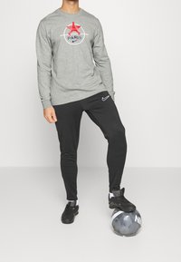 Graues Langarmshirt mit rotem und blauem grafischen "PARIS"-Logo, kombiniert mit schwarzen tapered Sporthosen und schwarzen Sportschuhen, stehend auf einem Fußball.