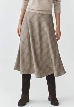 Personne portant une jupe midi beige à carreaux en ligne A, un pull beige et des bottes hautes marron foncé, debout devant un fond uni.