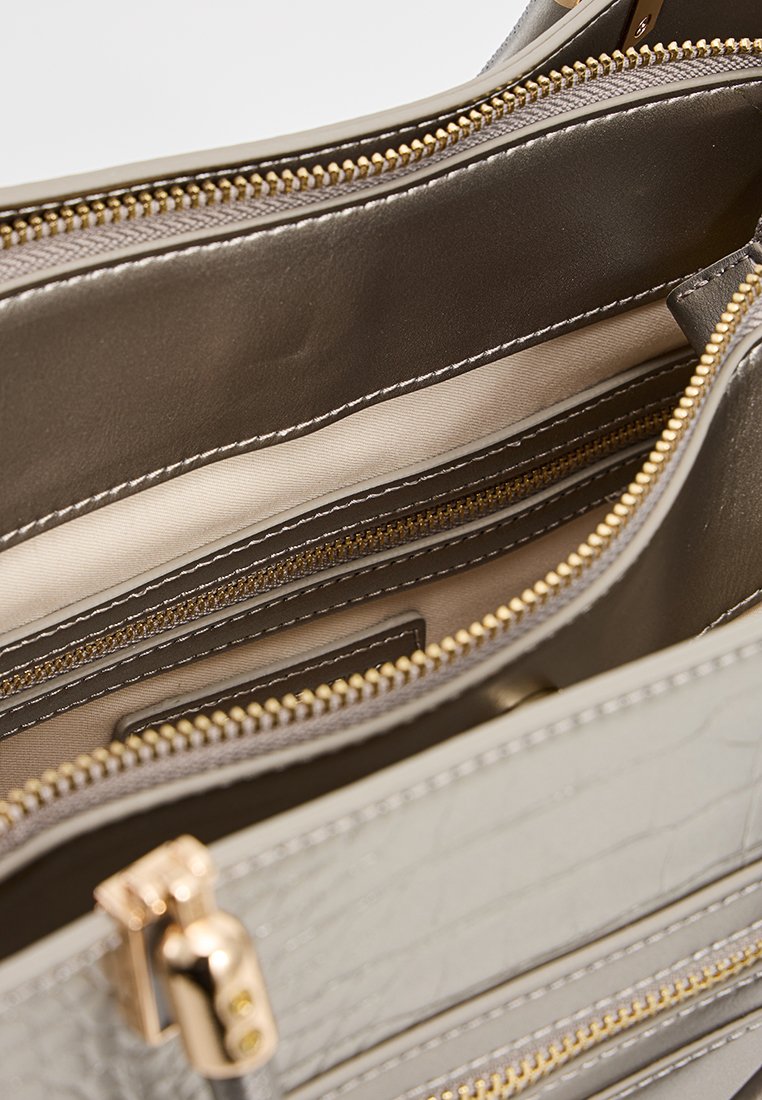Intérieur d'un sac à main structuré gris, avec de multiples compartiments dotés de fermetures éclair dorées et d'une doublure beige, mettant en valeur une surface lisse et texturée.