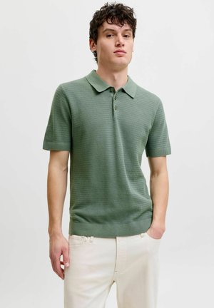 Jeune homme aux cheveux bouclés et foncés portant un polo texturé vert sauge à manches courtes et un pantalon beige clair, debout avec une main dans la poche.