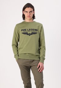 Maslinastozeleni dukserica s crnim natpisom "PME LEGEND AMERICAN CLASSIC" i dizajnom krila. Ima rebraste manžete i donji rub. Uparen s kaki hlačama.