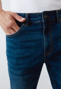 Jeans blu scuro in denim, con cuciture arancioni, una tasca frontale e chiusura con bottone in vita. Vista ravvicinata sull'area della vita.