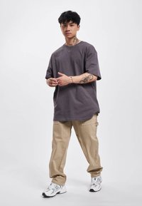 Grå oversized t-shirt, beige cargo bukser og hvide sneakers med grå accenter. Modellen står foran en simpel hvid baggrund.