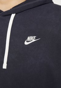 Marinblå hoodie med vita dragsnören; har en broderad vit Nike-logga på bröstet; tyget verkar mjukt och något texturerat.