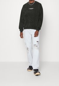 Czarna, oversize'owa bluza z logo, jasnoniebieskie jeansy z przetarciami oraz czarne sneakersy z fakturowaną podeszwą. Prosty łańcuszek jako dodatek.