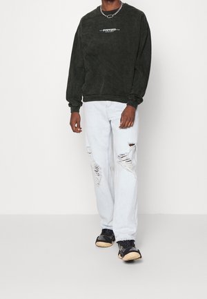 Sudadera negra de gran tamaño con logo, vaqueros azules claros desgastados y zapatillas negras con suelas texturizadas. Collar sencillo de cadena como detalle.