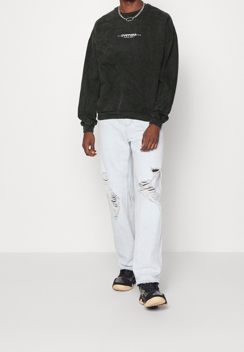 Czarna, oversize'owa bluza z logo, jasnoniebieskie jeansy z przetarciami oraz czarne sneakersy z fakturowaną podeszwą. Prosty łańcuszek jako dodatek.