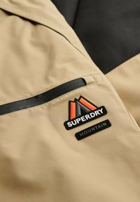 Beżowa kieszeń kurtki z czarnym zamkiem błyskawicznym i naszywką z logo "Superdry Mountain" z pomarańczowym i szarym wzorem górskim.