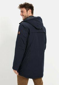 Marineblaue wasserdichte Jacke mit Kapuze, aus strukturiertem Stoff, mit verstellbaren Säumen und einem Logo-Patch am Ärmel.