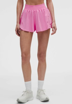 Mujer con pantalones cortos deportivos rosas, calcetines blancos y zapatillas blancas, de pie sobre un fondo liso.