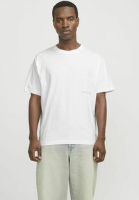 Weißes Baumwoll-T-Shirt mit lockerer Passform, Rundhalsausschnitt und kleinem roten Textdetail an der Vorderseite in der Nähe des Saums. Getragen mit heller Denim.