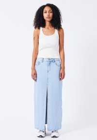 Falda larga de mezclilla en azul claro con abertura frontal, combinada con una camiseta ajustada blanca. La modelo lleva zapatillas deportivas blancas. Textura suave.